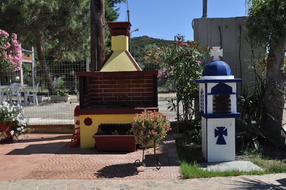 A serene outdoor patio features a colorful brick barbecue surrounded by vibrant flowers, a miniature blue-and-white chapel-like structure, and lush greenery, ideal for a relaxing vacation retreat.