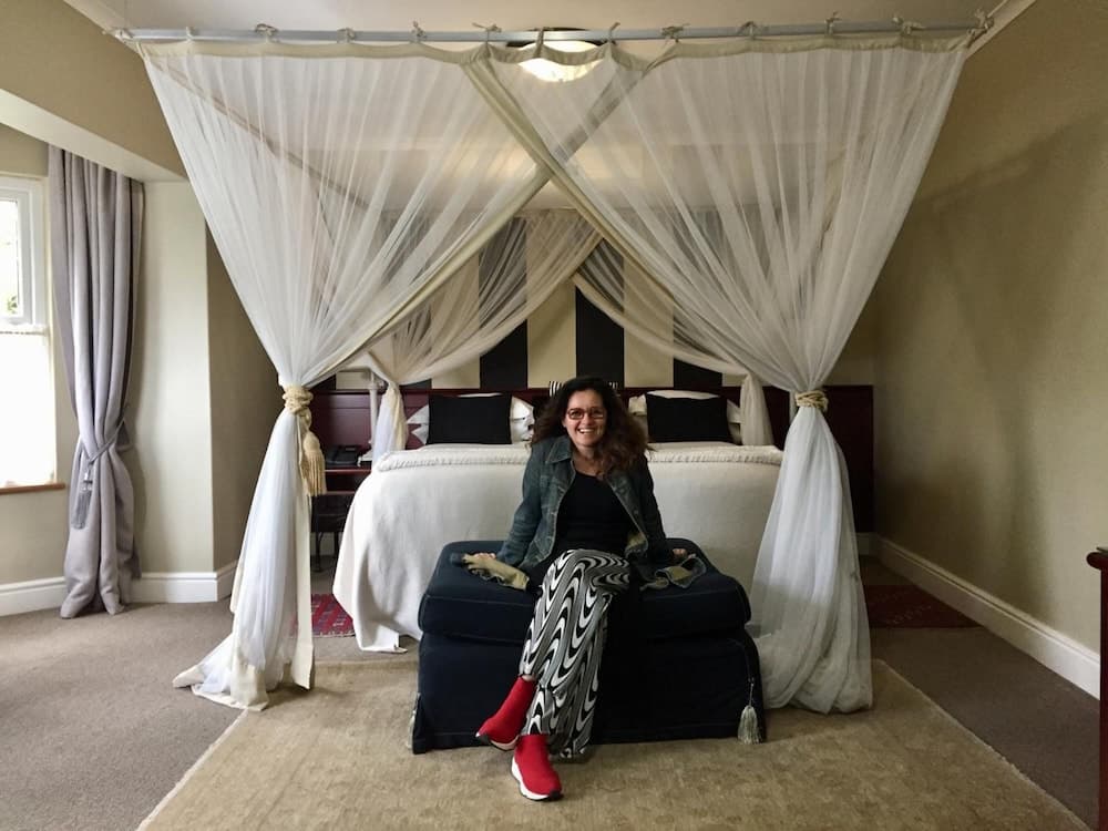 A cheerful traveler sits on a suitcase in a cozy hotel room featuring a canopy bed draped with sheer curtains, ideal for a relaxing getaway.
