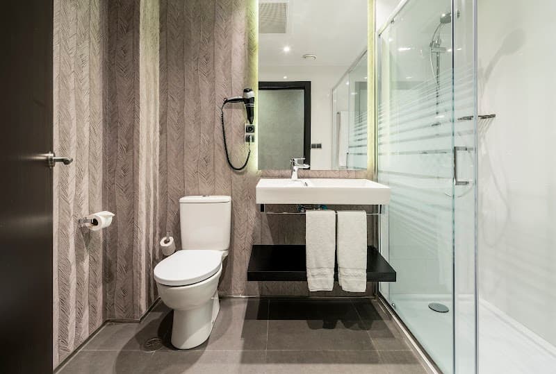 A modern hotel bathroom in Barcelona features sleek black and white decor, a glass-enclosed shower, and a wall-mounted sink, ideal for travelers seeking comfort and style during their stay.