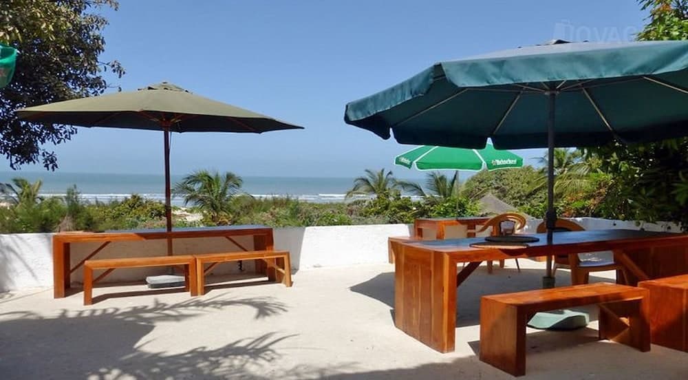 A serene beachside patio features wooden tables under large umbrellas, surrounded by lush greenery and offering a tranquil view of the ocean, perfect for relaxing getaways or coastal dining experiences.