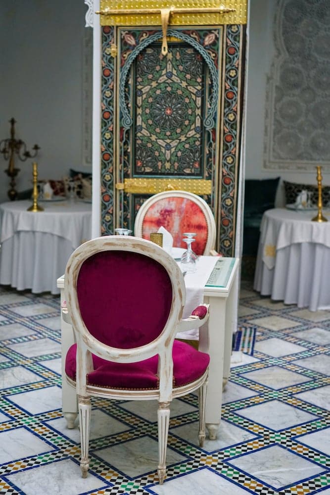 An elegantly decorated dining area features a vibrant magenta chair and ornate Moroccan-style decor, creating an inviting atmosphere ideal for luxury travel experiences.