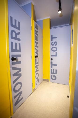 A vibrant indoor corridor with bold yellow and white doors labeled "NOWHERE," "SOMEWHERE," and "GET LOST" invites curiosity and exploration, perfect for a unique museum or art gallery experience.