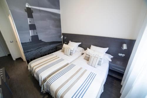 A cozy hotel room features two neatly made beds with striped linens, a lighthouse mural on the wall, and modern furnishings, ideal for travelers seeking comfort and a maritime ambiance.