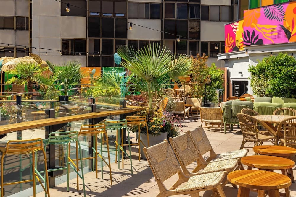 A vibrant rooftop terrace adorned with colorful chairs, lush greenery, and a striking mural creates an inviting urban oasis perfect for leisurely dining or social gatherings.