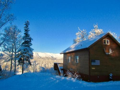 A cozy wooden cabin adorned with an American flag nestles in a serene snowy landscape under a clear blue sky, ideal for winter retreats and nature lovers seeking tranquility.