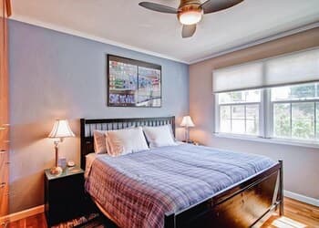 A cozy bedroom with soft blue walls, a neatly made bed adorned with a plaid blanket, and natural light streaming through large windows, creates an inviting atmosphere ideal for travelers seeking comfort and relaxation.