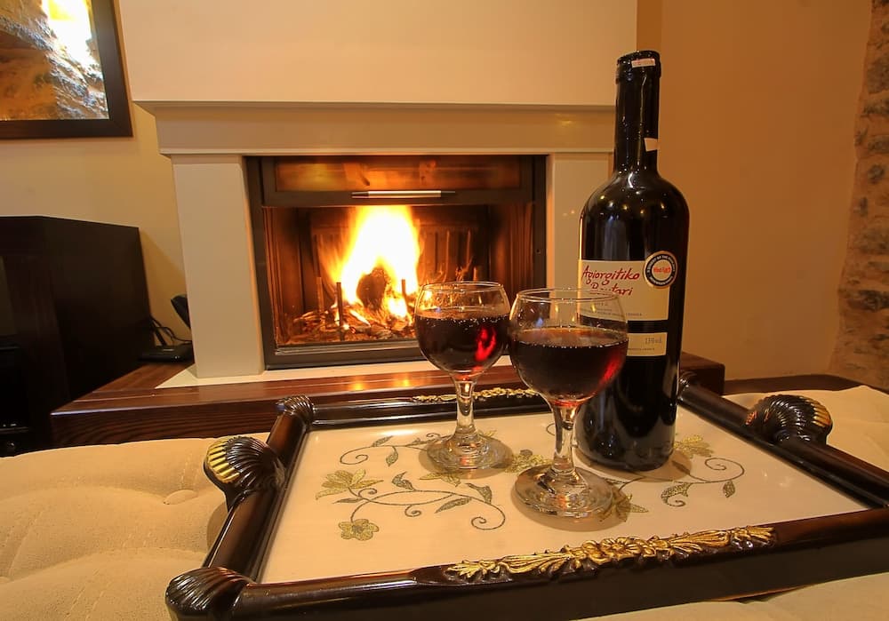 A cozy indoor setting features a warm fireplace, a tray with two glasses of red wine and a bottle labeled "Agiorgitiko," creating an inviting atmosphere perfect for a romantic getaway or a relaxing evening in.