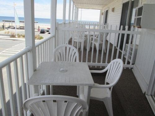 A serene beachside porch features white plastic chairs and tables under a sunny sky, ideal for relaxing oceanfront getaways.