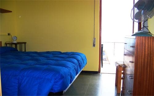 A cozy bedroom with a vibrant yellow wall, blue bedding, wooden dresser, and balcony view offers a serene retreat for travelers seeking simplicity and comfort.