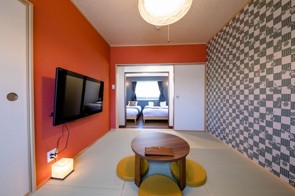 A cozy hotel room features vibrant orange walls, a patterned wallpaper, a low wooden table with cushions, a wall-mounted TV, and a view into a bedroom area, ideal for travelers seeking comfort and modern amenities in a compact space.