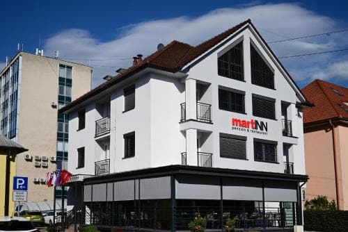 A charming white building with red-tiled roofs houses the martINN hotel and restaurant under a bright blue sky, offering travelers a cozy urban retreat with balconies and modern amenities.