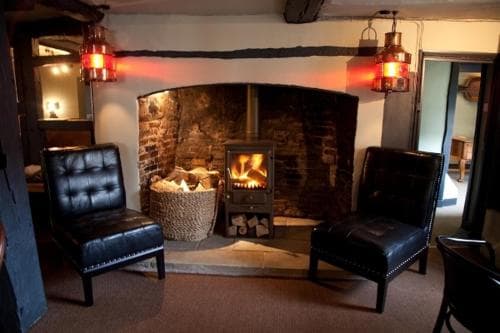 A cozy indoor lounge features a rustic stone fireplace with a glowing wood-burning stove, flanked by leather armchairs under warm lantern lighting, ideal for travelers seeking relaxation in a traditional pub setting.