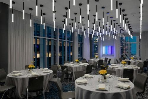 An elegant banquet hall with round tables draped in white linens, modern hanging lights, and large windows offering a cityscape view, creates an upscale dining atmosphere ideal for corporate events or luxurious gatherings.