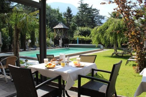 A serene outdoor dining area by a lush green lawn and poolside, featuring breakfast tables set under a glass roof, surrounded by trees and distant mountains, ideal for a relaxing vacation retreat.