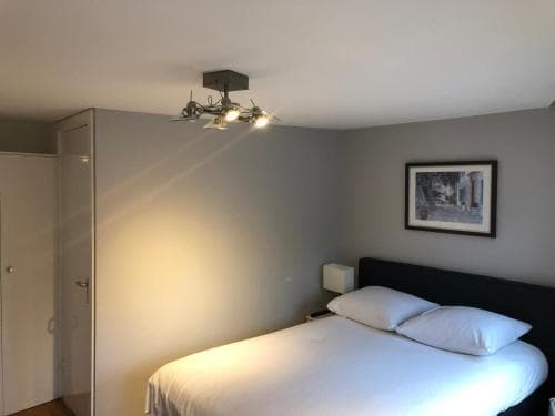 A cozy hotel room features a neatly made bed under warm ceiling lights, framed artwork on the wall, and minimalist decor, ideal for travelers seeking comfort and simplicity.