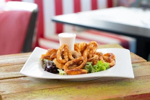 A plate of crispy calamari garnished with fresh greens sits invitingly on a rustic wooden table in a vibrant outdoor café setting, perfect for travelers seeking a casual dining experience.