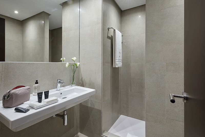 A serene and modern bathroom in the Hotel Rey Alfonso X Sevilla features sleek beige tiles, a minimalist sink setup with toiletries, a vase of white flowers, and a towel rack, ideal for travelers seeking comfort and elegance during their stay.