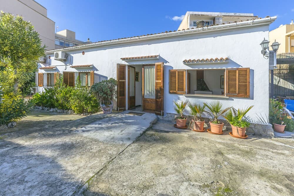 A charming white Mediterranean-style house with wooden shutters, potted plants, and lush greenery exudes a serene and inviting atmosphere, perfect for a relaxing vacation getaway.