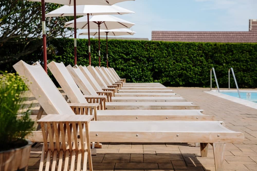 A serene poolside scene features neatly arranged wooden lounge chairs under white umbrellas, set against lush green hedges, ideal for relaxing vacations or leisurely getaways.