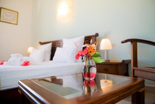 A cozy hotel room exudes warmth with its neatly made bed adorned with red flowers, a glass-topped coffee table featuring vibrant blooms, and soft lighting, ideal for travelers seeking relaxation and comfort.