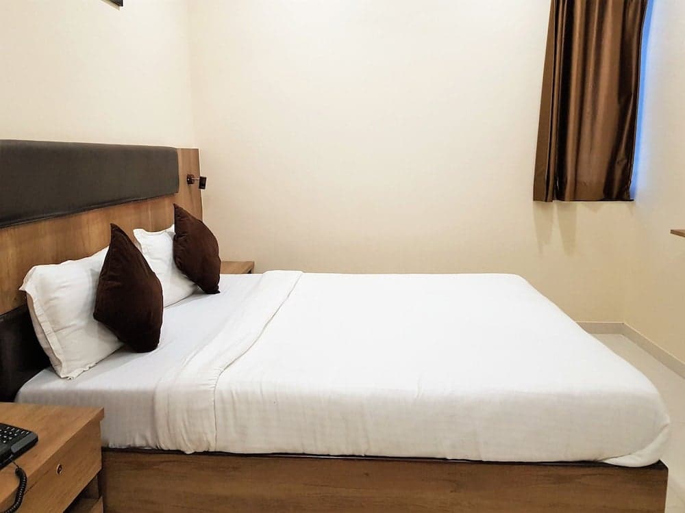 A cozy hotel room features a neatly made bed with white linens and brown pillows, a wooden bedside table with a phone, and a curtained window, ideal for travelers seeking comfort and simplicity.