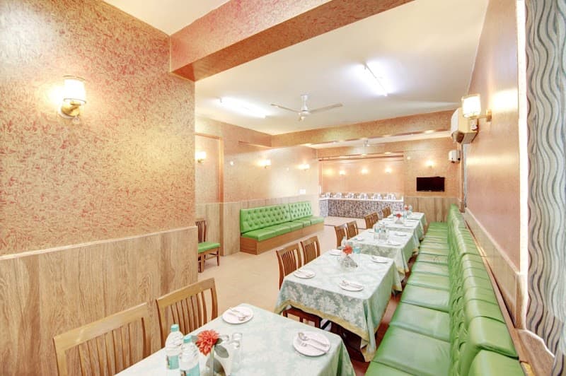 A cozy indoor dining area at a hotel in Noida, featuring green upholstered benches, wooden chairs, and tables set with floral-patterned tablecloths, ideal for travelers seeking a comfortable meal spot near the Amazon office.