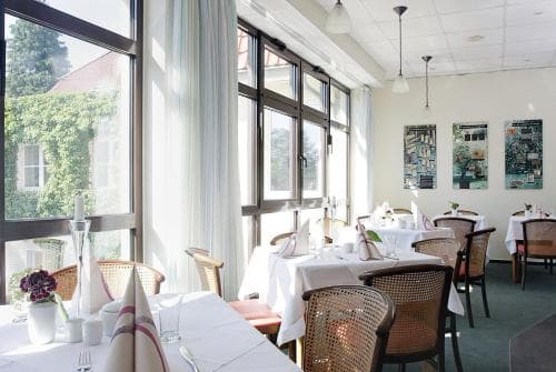 A cozy restaurant interior bathed in natural light from large windows, featuring neatly arranged tables with white linens, wicker chairs, and vibrant artwork on the walls, ideal for a serene dining experience or a romantic getaway.