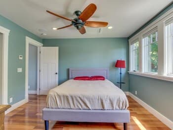 A cozy bedroom with light blue walls, wooden flooring, and a neatly made bed under a ceiling fan, featuring natural light from large windows and a red lamp, ideal for travelers seeking a serene and comfortable stay.