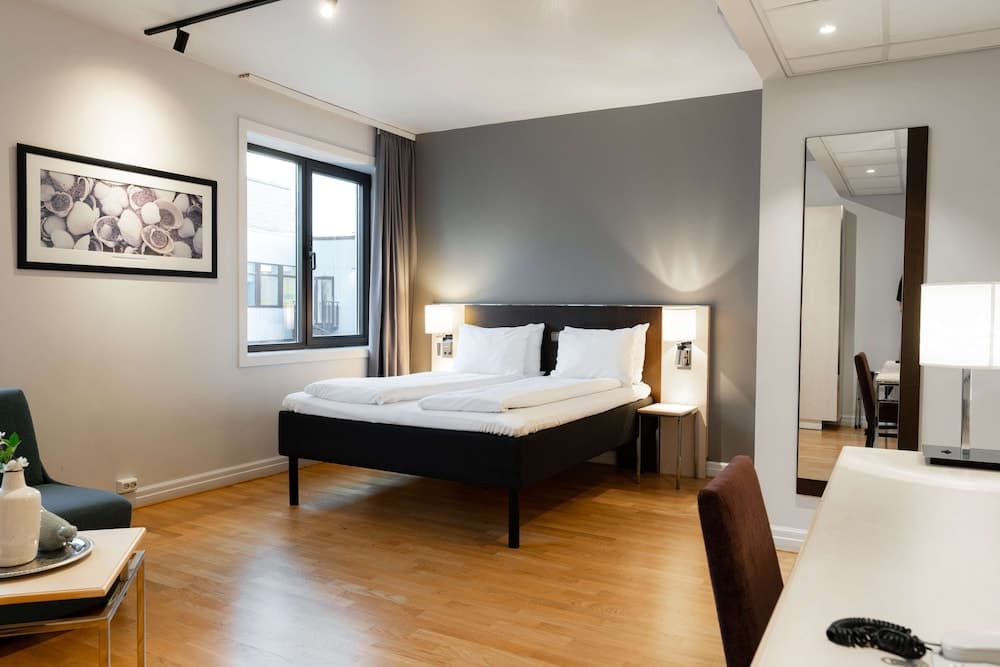 A cozy hotel room features a neatly made bed with white linens, wooden flooring, modern lighting, and a large mirror, creating a serene atmosphere ideal for travelers seeking comfort and style.