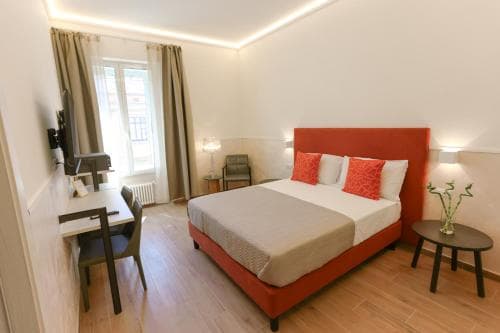 A cozy hotel room features a vibrant red bed with white linens, a wooden desk by the window, and warm lighting, creating an inviting atmosphere ideal for travelers seeking comfort and relaxation.