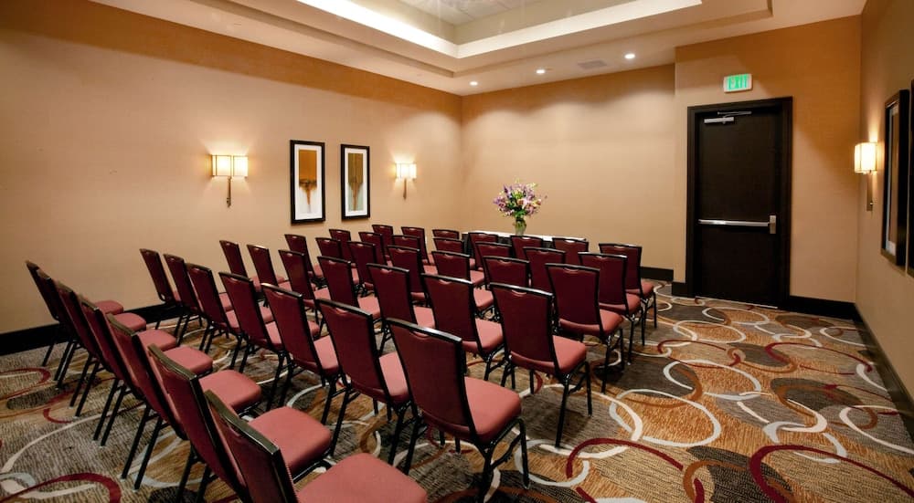 A cozy conference room with neatly arranged red chairs, patterned carpeting, framed artwork, and soft lighting creates an inviting atmosphere ideal for business meetings or small gatherings.