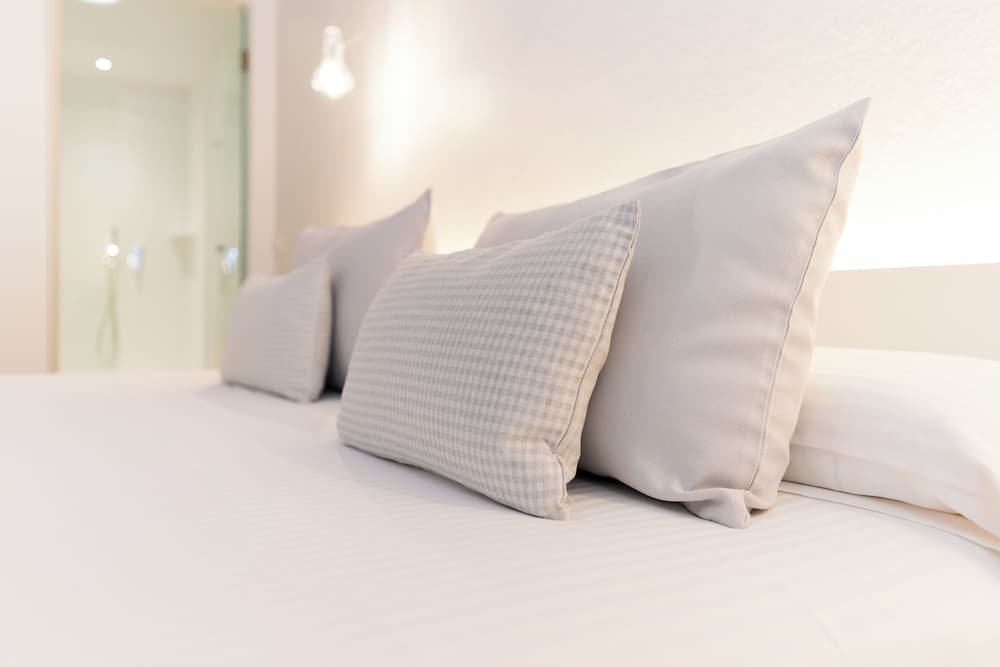 A serene hotel bedroom scene features a neatly arranged bed with white linens and gray pillows, complemented by soft lighting and a visible bathroom entrance, ideal for travelers seeking comfort and relaxation.