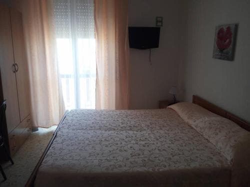 A cozy hotel room features a neatly made bed with patterned bedding, soft pink curtains framing a bright window, a mounted TV, and minimalist decor, ideal for travelers seeking comfort and simplicity.