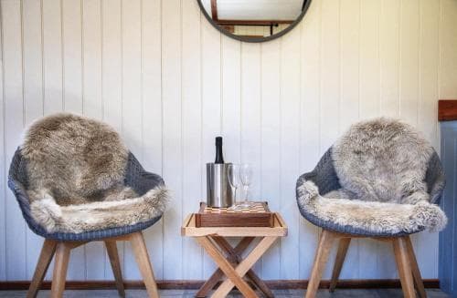 A cozy cabin interior features two wooden chairs draped with plush fur throws, flanking a small table with a wine bottle and glasses, evoking a serene and inviting atmosphere perfect for a relaxing getaway.