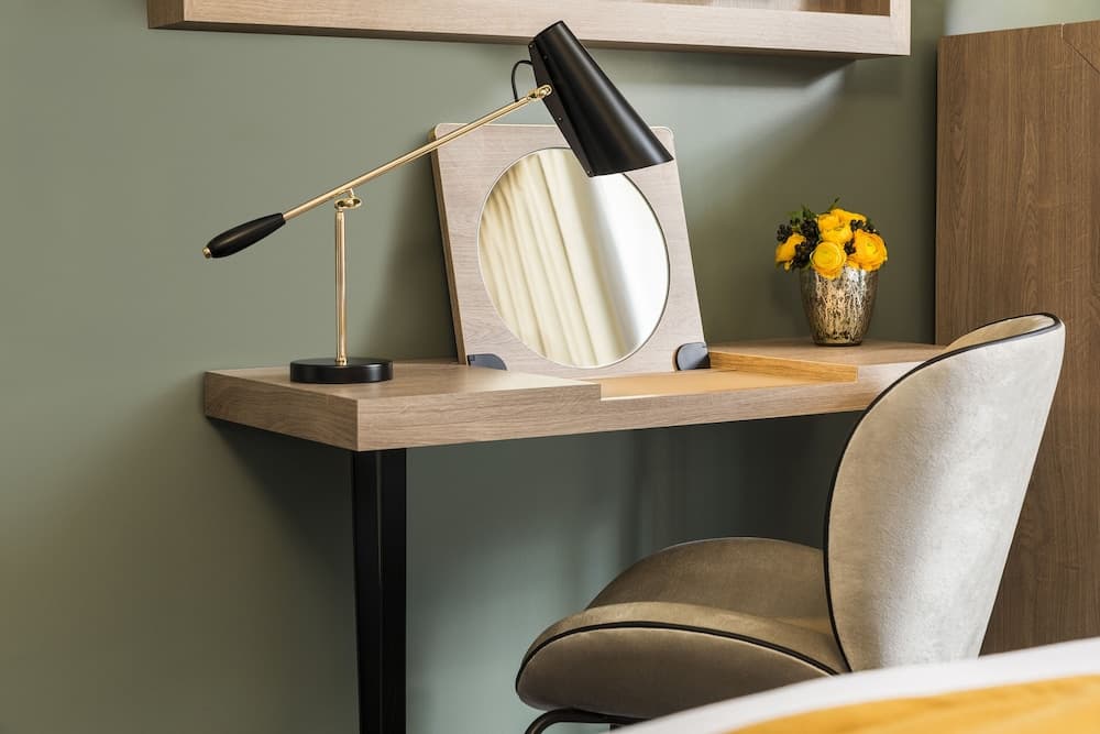 A cozy hotel room corner features a sleek wooden desk with a stylish lamp, round mirror, and vibrant yellow flowers in a vase, creating an inviting workspace perfect for travelers seeking comfort and functionality.