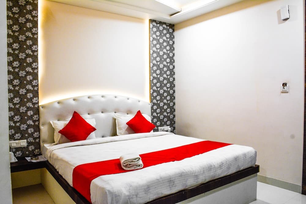 A cozy hotel room features a neatly made bed with white linens, red accents, and a tufted headboard, set against patterned wallpaper, ideal for travelers seeking comfort and simplicity.