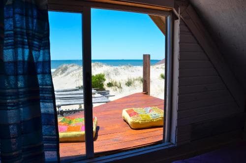 A cozy attic window frames a serene beach view with vibrant cushions on a wooden deck, perfect for a tranquil seaside getaway.
