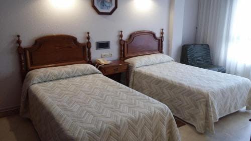 A cozy hotel room features two neatly made single beds with wooden headboards, a small nightstand, and soft natural light filtering through sheer curtains, ideal for travelers seeking a comfortable and tranquil stay.