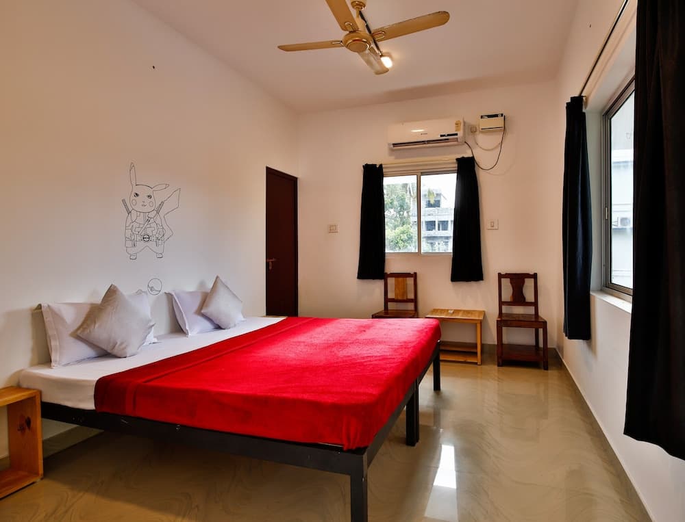 A cozy hotel room features a bed with a vibrant red blanket, wooden furniture, black curtains, and a wall mural, creating a welcoming atmosphere ideal for travelers seeking comfort and simplicity.