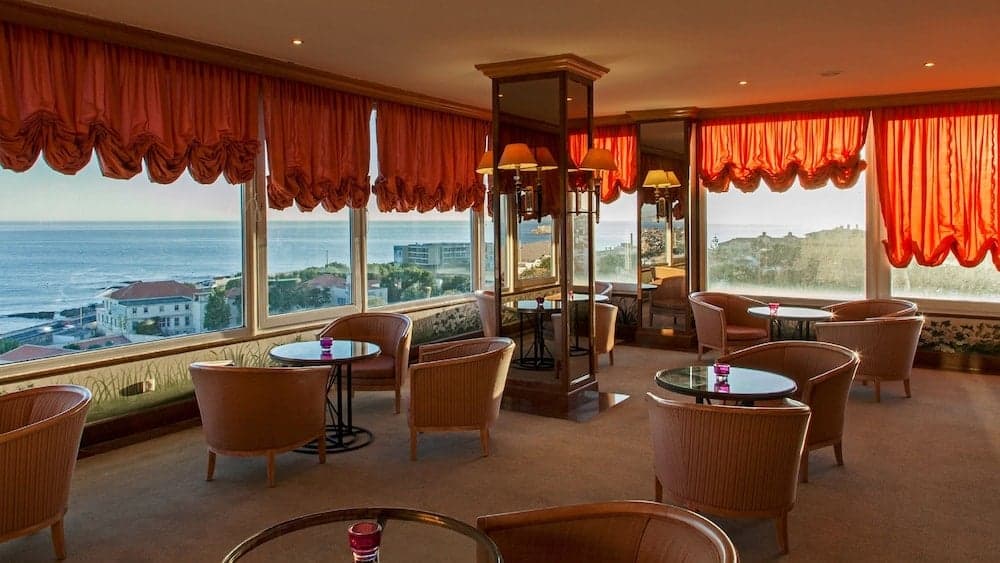 A cozy lounge with plush armchairs and tables offers a serene seaside view through large windows adorned with elegant red curtains, ideal for travelers seeking relaxation in a luxurious coastal retreat.