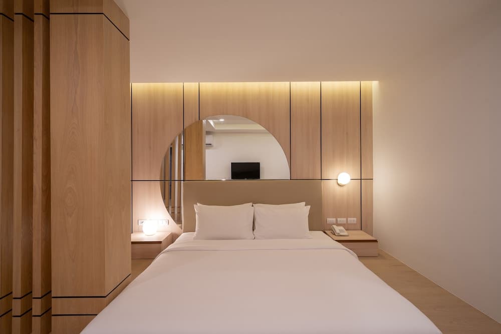 A serene hotel room features a cozy bed with white linens, wooden accents, soft lighting, and a unique arched mirror, ideal for travelers seeking a tranquil and stylish accommodation experience.