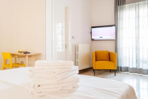 A cozy hotel room features a neatly made bed with folded towels, a vibrant yellow armchair, a desk with a chair, and a wall-mounted TV, creating a welcoming atmosphere ideal for travelers seeking comfort and relaxation.