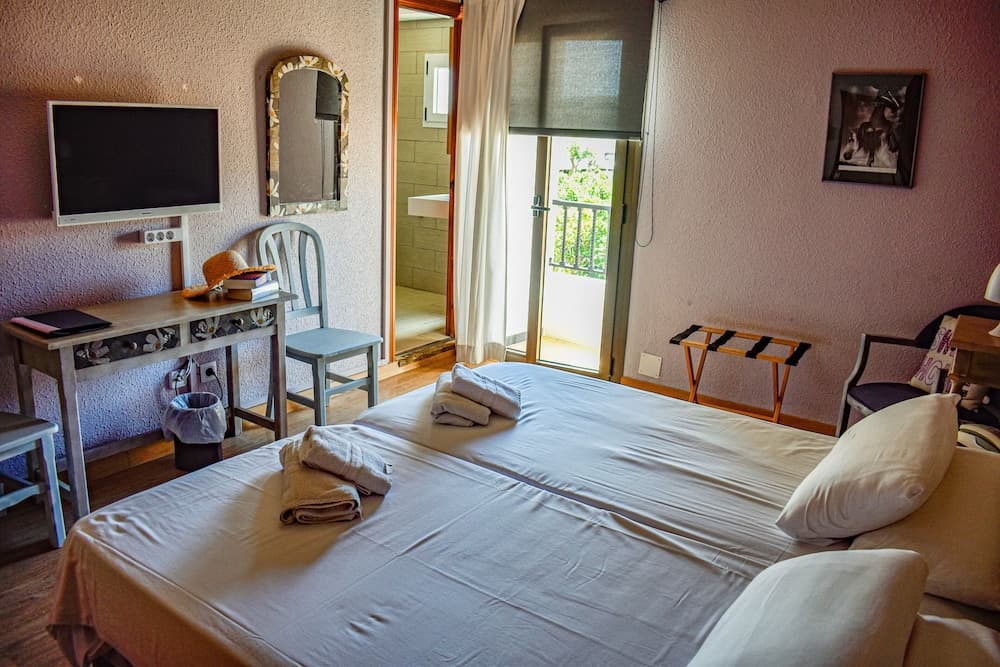 A cozy hotel room with soft pink walls, a neatly made bed, a desk with a TV, and a balcony door inviting natural light, perfect for travelers seeking comfort and relaxation.