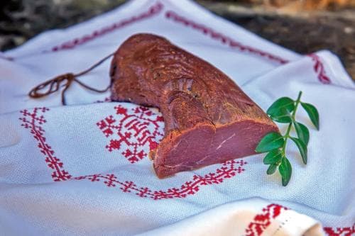 A rustic slice of cured meat rests on an embroidered cloth adorned with red patterns, evoking a traditional culinary scene perfect for cultural food tourism.
