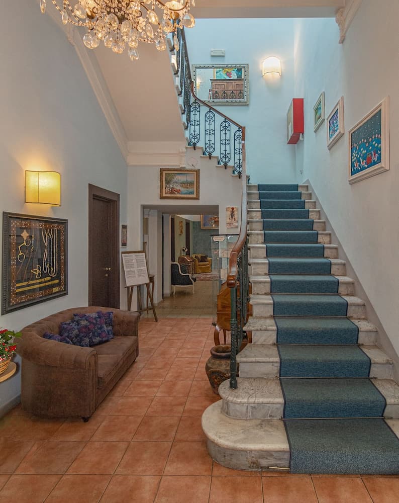 A cozy hotel lobby features a grand staircase with blue carpeting, ornate railings, and warm lighting, creating an inviting atmosphere perfect for travelers seeking comfort and elegance.