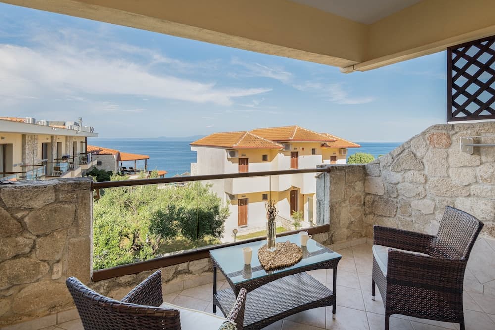 A serene coastal balcony offers a picturesque view of the sea and charming buildings, featuring wicker furniture and a stone railing, ideal for a relaxing vacation getaway.