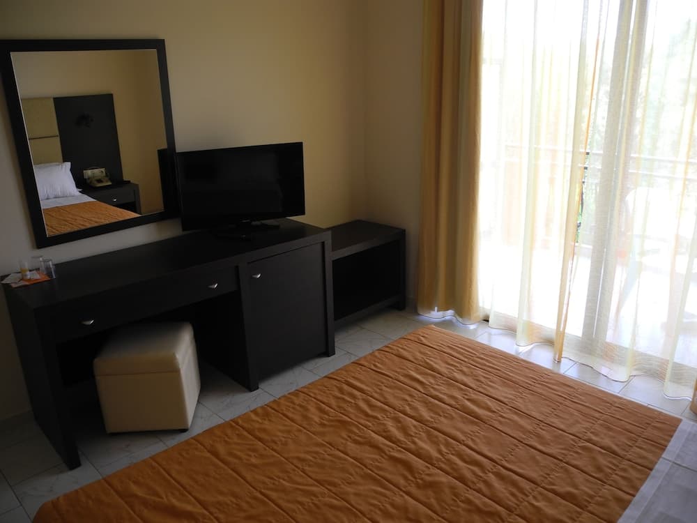 A cozy hotel room features a neatly made bed, a sleek black desk with a TV and mirror, soft yellow curtains framing a balcony view, ideal for travelers seeking comfort and relaxation.