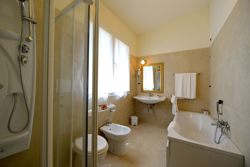 A cozy hotel bathroom features beige tiles, a bathtub with a handheld shower, a bidet, a sink with a gold-framed mirror, and soft natural light filtering through sheer curtains, ideal for travelers seeking comfort and cleanliness.