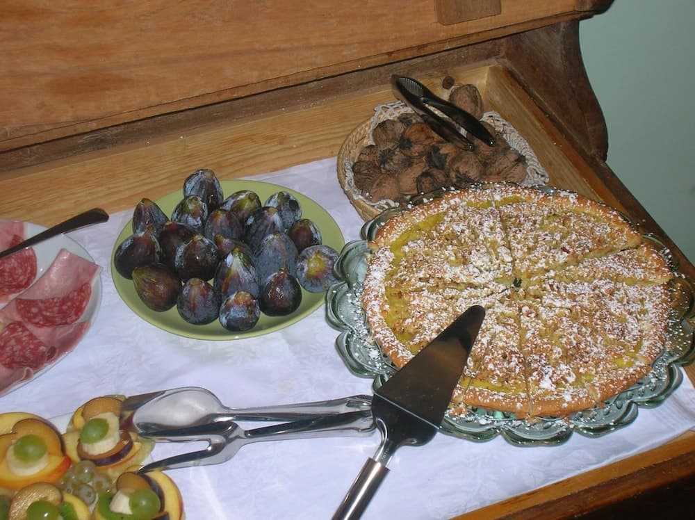 A cozy indoor buffet table displays an inviting spread of fresh figs, a powdered sugar-topped pie, assorted meats, and fruit slices, perfect for a rustic culinary exploration or a charming bed-and-breakfast experience.