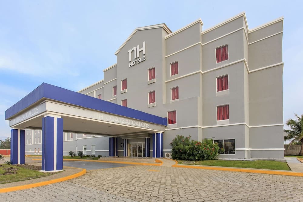 A modern NH hotel with a striking blue and gray facade welcomes travelers with its spacious driveway and lush greenery, ideal for business or leisure stays in a serene urban setting.